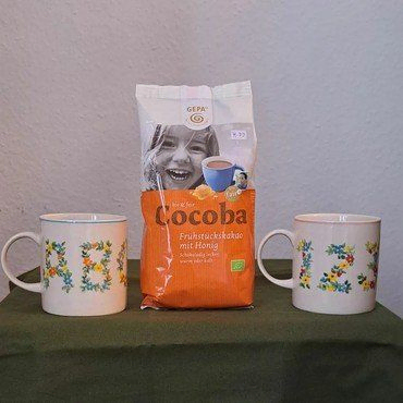 Cocoba Kakaopackung zwischen zwei Tassen mit floralen Buchstaben-Designs.